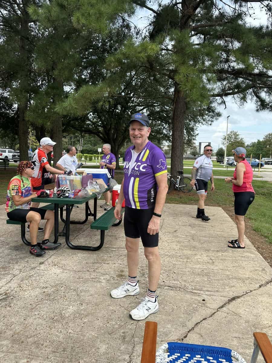 Zube Volunteer Salute Smiling cyclist in purple NWCC jersey at community bike event in park.